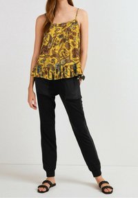 Haut jaune à motifs avec des volants au bas, associé à un pantalon noir fuselé. Le mannequin porte des sandales noires avec des brides tressées.