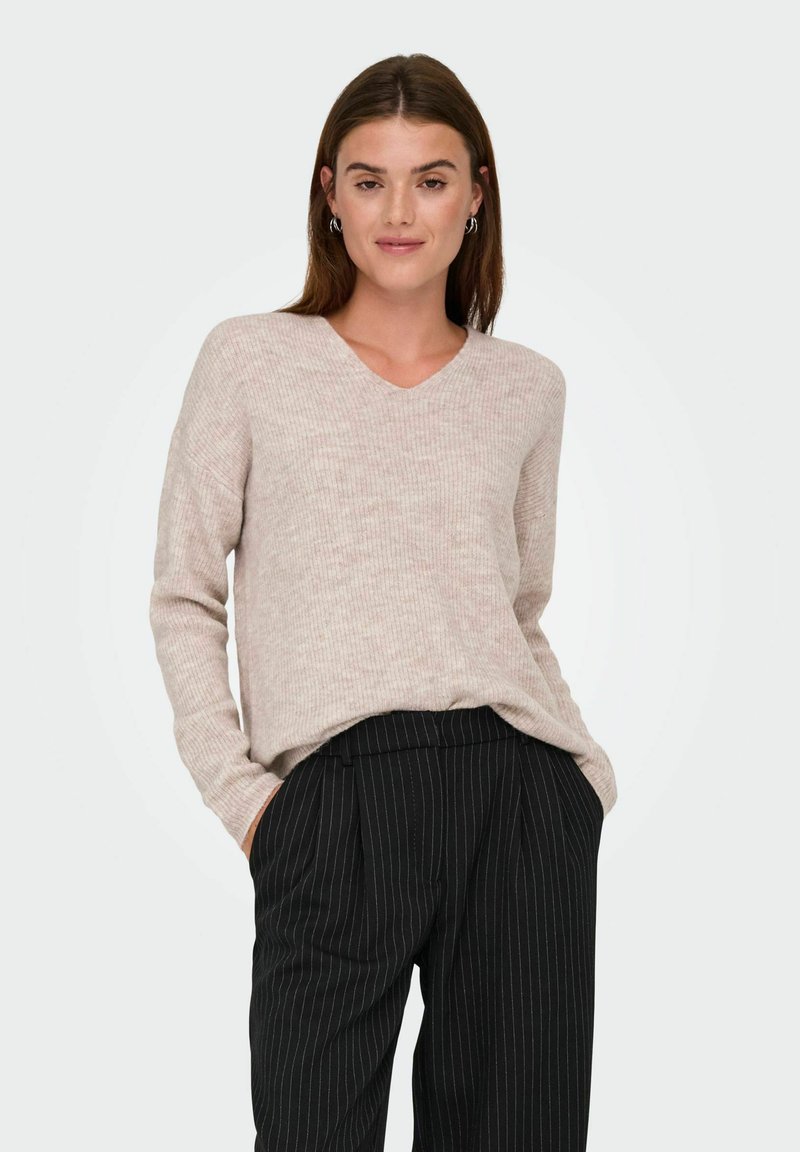 Femme aux cheveux bruns portant un pull en maille beige et un pantalon noir à fines rayures, les mains dans les poches, debout devant un fond uni.