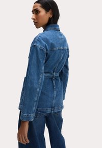 Giacca di denim in blu medio con vita cinturata, maniche lunghe e colletto classico. Presenta cuciture visibili e una vestibilità rilassata.