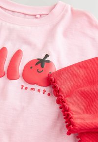 Růžová bavlněná košile s grafikou rajčete, červenými doplňky a volánovými manžetami na rukávech. Design obsahuje černé detaily a nápis "rajče".