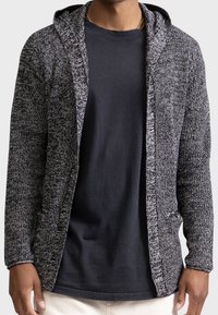 Hombre vestido con una camiseta negra y un cárdigan gris de punto con capucha, de pie contra un fondo claro.