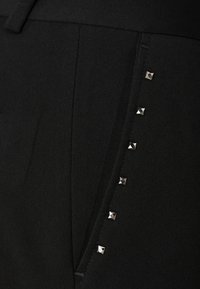 Zwarte broek van een textuurstof, met zijzakken en een reeks zilveren piramidestuds langs de naad van de zak voor extra detail.