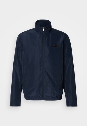 Námornícky bomber jacket s vystuženým golierom, predným zipsom a elastickým pásom; obsahuje malý čierny logový znak na hrudi. Hladká textúra.