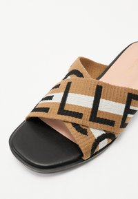 Sandale à bande croisée marron et crème avec semelle en caoutchouc noire ; présente des lettres en gras et une texture tissée sur le dessus.