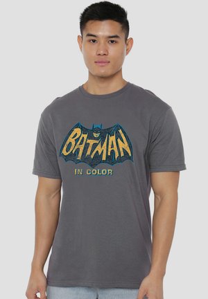 Junger Mann trägt ein graues T-Shirt mit einem Vintage-Batman-Logo in Gelb und Blau und dem Text "IN COLOR" auf der Vorderseite.
