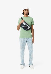 Groene katoenen t-shirt, blauwe gebloemde jeans en een zwarte nylon schoudertas met witte en teal accenten; model draagt een witte pet en sneakers.