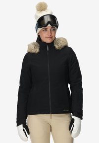Mujer con chaqueta de invierno negra con borde de piel, pantalones beige, mitones blancos, gorro blanco con pompón y gafas de esquí negras en la frente.