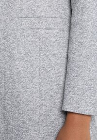 Grå stickad tröja med en mjuk textur, långa ärmar och en diskret bröstficka. Tyget har ett subtilt melerat mönster.