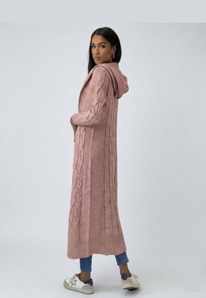 Vrouw met een lange roze kabelgebreide capuchoncardigan, blauwe spijkerbroek en witte sneakers, kijkt over haar schouder tegen een effen achtergrond.