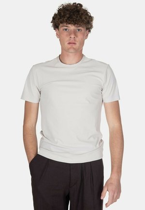 Giovane uomo con capelli ricci che indossa una camicia beige chiaro a maniche corte e pantaloni scuri, in piedi con una mano in tasca davanti a uno sfondo semplice.