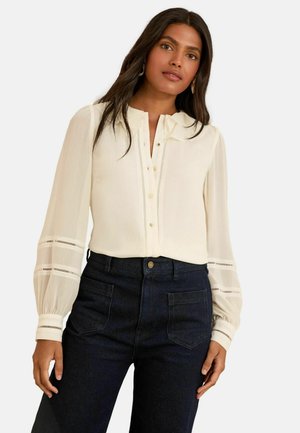 Weiße Bluse mit langen, gepufften Ärmeln, die mit Spitzenakzenten und einer Knopfleiste versehen ist. Kombiniert mit dunklen, hochtaillierten Jeanshosen.