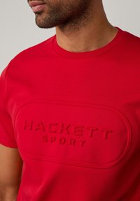 Camiseta de algodón roja con un logotipo texturizado y en relieve que dice "HACKETT SPORT" en un diseño rectangular en la parte frontal.