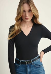 Czarny bodysuit z długim rękawem i dekoltem w kształcie litery V, w połączeniu z niebieskimi dżinsami o wysokim stanie. Stylizacja zawiera czarny pasek z złotymi akcentami.