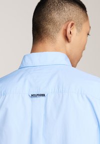 Light blue collared shirt with a smooth fabric. Features a black "HILFIGER" label at the back neck and a structured yoke.