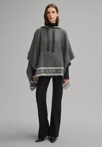 Poncho grigio con cappuccio, dotato di lacci neri, orlo a righe con dettaglio del logo e bordi sfrangiati. Abbinato a pantaloni neri a zampa.
