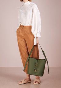 Žena v bílé pruhované blůze a hnědých kalhotách, držící velkou zelenou kabelku a obutá v béžových sandálech na otevřené špičce.