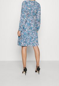 Langärmliges Kleid mit blauem Blumenmuster, aus leichtem Stoff, das oberhalb des Knies endet. Kombiniert mit schwarzen Absatzschuhen.