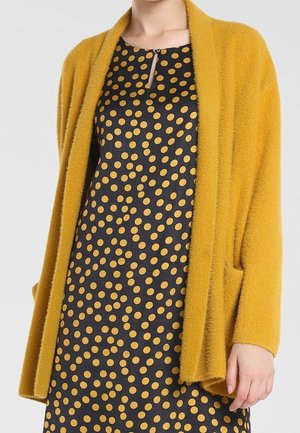 Vrouw draagt een zwarte jurk met mosterdkleurige polkadots en een mosterdkleurige, zachte open cardigan met zakken.