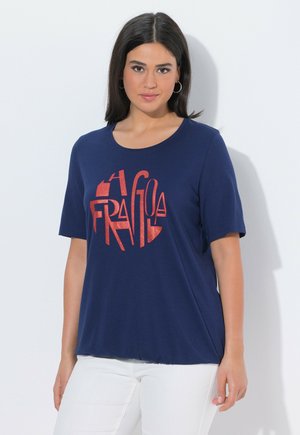 Kvinde med mørkt hår iført navyblå T-shirt med rødt grafisk tekst og hvide bukser, stående mod en enkel lys baggrund.