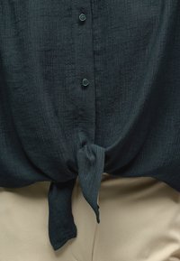 Chemise texturée foncée à boutons, nouée en un nœud à la taille, portée sur un pantalon beige clair.