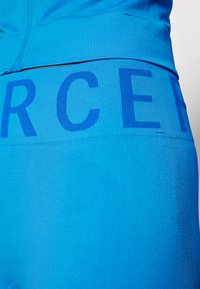 Modre atletske legice z teksturiranim pasom, na katerem je v debeli pisavi napisano "FORCE", gladka tkanina z rahlim sijajem in zasnovane konture.