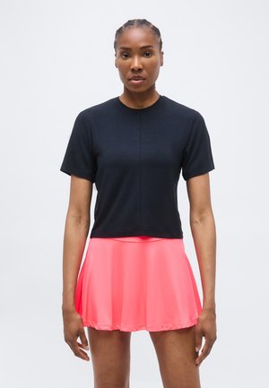 Žena stoji noseći crni top s kratkim rukavima i svijetlo ružičastu suknju na volane na jednostavnoj svijetloj pozadini.