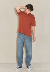 Koszulka z krótkim rękawem w kolorze rdzawego rdzawo-złotym, zestawiona z jasnoniebieskimi jeansami o prostych nogawkach oraz czarnymi niskimi sneakersami. Materiał o wyraźnej teksturze i swobodny krój.