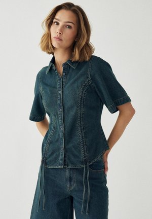 Žena s vlasmi po plecia, ktorá má na sebe priliehavú krátkym rukávom zapínanú rifľovú košeľu a zodpovedajúce rifľové nohavice na jednoduchom pozadí.