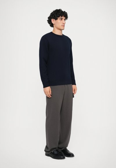 Námořnický pletený svetr s kulatým výstřihem, kombinovaný se šedými širokými kalhotami. Černé boty s minimalistickým designem dotvářejí outfit.