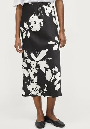 Jupe noire à motifs floraux blancs, à la texture lisse, longueur cheville, et silhouette ajustée. Portée avec des chaussures plates noires.