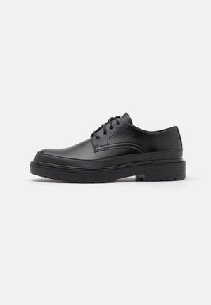 Chaussure habillée pour homme en cuir noir à lacets, avec une semelle épaisse en caoutchouc et un petit talon, présentée en profil sur fond blanc.