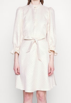 Femme portant une robe blanche texturée longueur genou avec un col haut à volants, des manches trois-quarts bouffantes et un nœud à la taille.