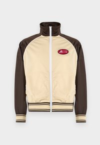 Beige en bruine zip-up jas met hoge kraag, gestreepte manchetten en zoom, met een ronde patch met een rood ontwerp op de linkerkant van de borst.