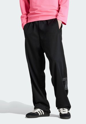 Osoba ubrana w czarne spodnie dresowe Adidas z bocznymi naszywkami z logo, różową bluzkę z długim rękawem oraz czarne buty sportowe Adidas z białymi paskami.