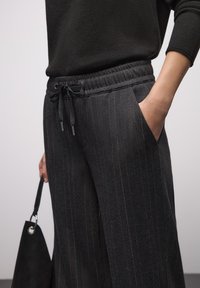 Mustat, ohutraitaiset housut, joissa on joustava vyötärönauha ja kiristysnaru. Housuissa on sivutaskut ja sileä pinta. Käsi pitelee mustaa laukkua.