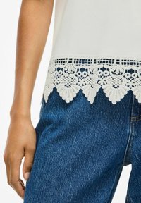Top blanco con un dobladillo de encaje que presenta patrones florales, combinado con jeans de mezclilla azul oscuro. La tela parece suave, con un acabado ligeramente texturizado.