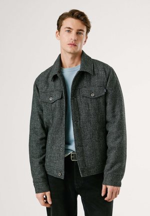 Chaqueta gris texturizada con dos bolsillos en el pecho, botones a presión y cuello, combinada con un suéter azul claro y pantalones negros.