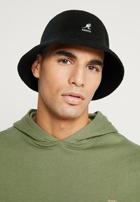 Cappello nero a secchiello realizzato in tessuto testurizzato, con logo Kangol sulla parte anteriore. Indossato con una felpa verde, che mostra una vestibilità comoda e un collo allentato.