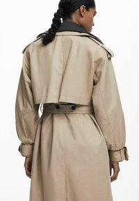 Manteau trench beige avec un design de dos court, col noir, taille cintrée et accents de bouton sur les manches. Texture lisse, style minimaliste.