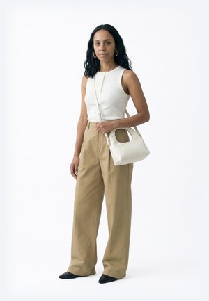 Femeie cu păr lung și negru, purtând un top alb fără mâneci, pantaloni largi bej, pantofi negri cu vârf ascuțit și având o geantă albă cu bretea de umăr.