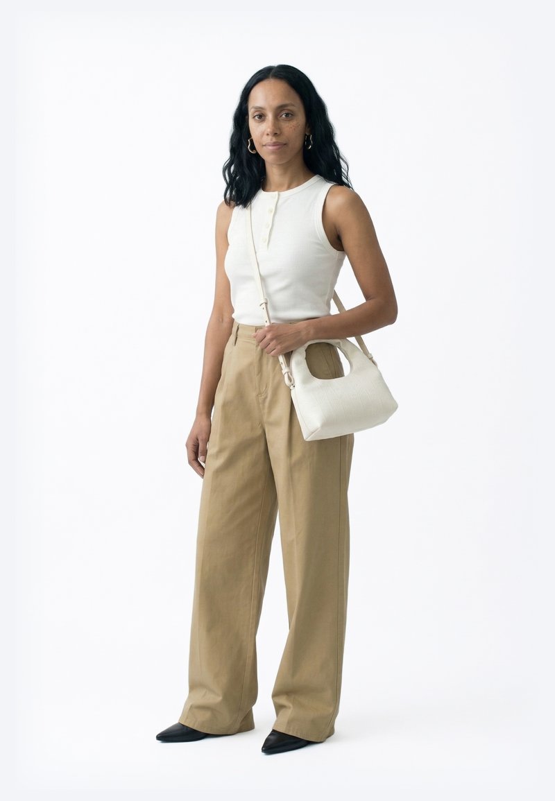 Pikkade mustade juustega naine, kes kannab valget varrukateta topi, beeže laiade pükstega, mustade teravate kingadega ning kannab valget õlakotiga käekotti.