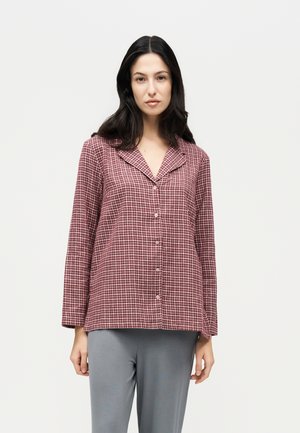Une femme porte une chemise boutonnée à carreaux bordeaux et blancs avec un pantalon gris, sur un fond clair et uni.