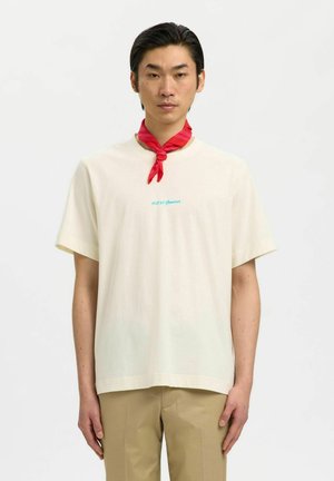 Jeune homme portant un t-shirt crème avec texte bleu, pantalon beige et un foulard rouge autour du cou, debout devant un fond blanc uni.