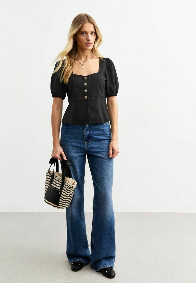 Zwarte top met pofmouwen, vierkante halslijn en knoopdetails, gecombineerd met wijde blauwe jeans. Vastgehouden wordt een gemêleerde geweven tas.