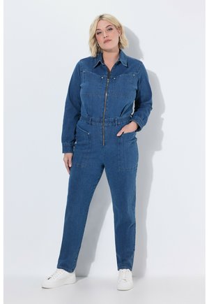 Frau mit kurzen blonden Haaren, die einen blauen Denim-Overall mit vorderem Reißverschluss und weißen Sneakern trägt, steht vor einem weißen Hintergrund.