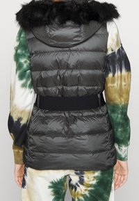 Svart uppblåsbar väst med en avtagbar fuskpälskrage och elastisk midja, bärs över en flerfärgad tie-dye-outfit.