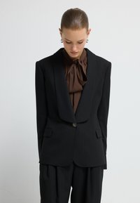 Zwarte gestreepte blazer met een geknipt revers, een enkele knoopsluiting en twee voorzakken, gecombineerd met een bruine strikblouse.