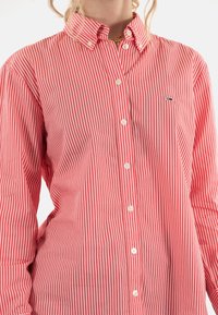 Camisa de botones a rayas rojas y blancas con cuello, que presenta botones blancos y un pequeño logo en el pecho. Textura de tela suave.