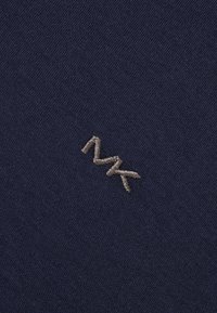 Jūras zila auduma virsma ar smalku, teksturētu austību, uz kuras ir izšūta bežā krāsā "MK" logo ar izliekta dizaina elementiem.