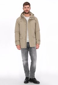 Chaqueta acolchada beige con capucha, con dos bolsillos delanteros, abrochada con botones, llevada sobre una camisa gris claro y jeans grises. Botas negras.